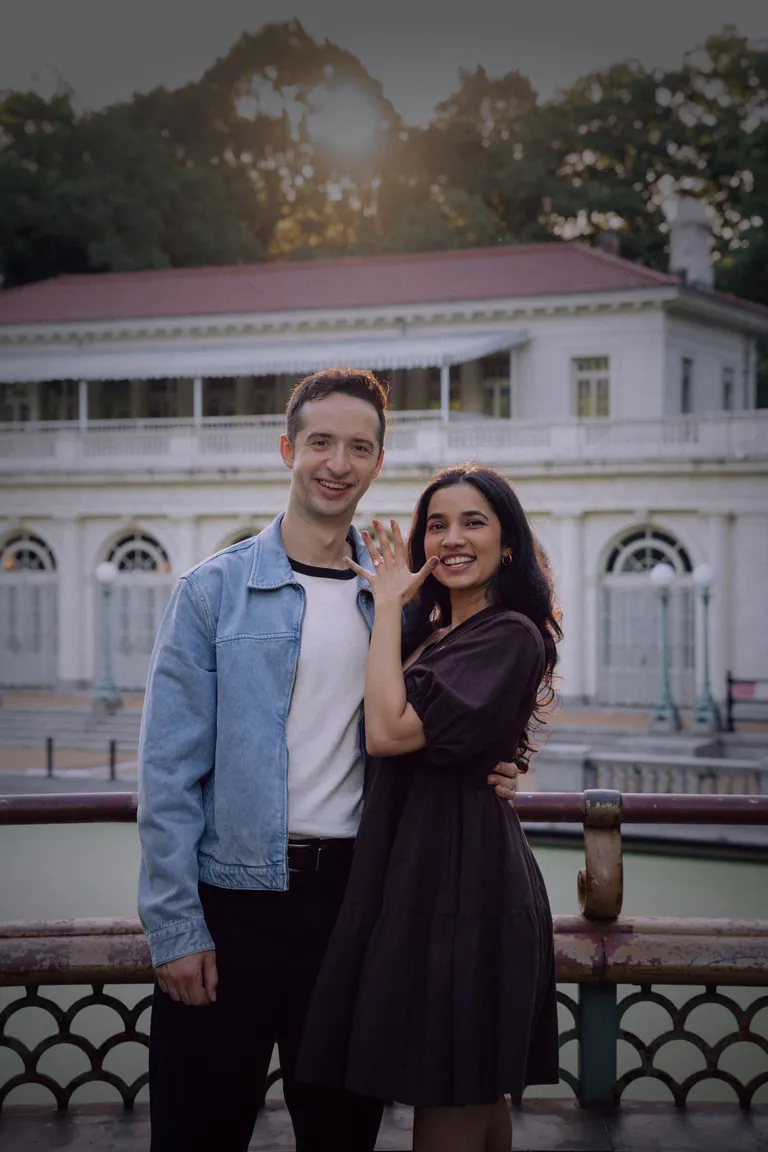 Caleb and Shruti smiling in front of the Prospect Park boathouse with Shruti showing her ring.